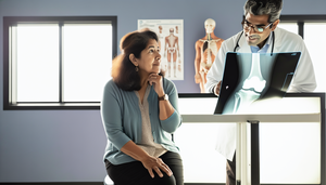 Mujer hispana de mediana edad en consultorio médico observando una radiografía de rodilla con signos de artrosis, como estrechamiento del espacio articular y osteofitos. Un médico hispano señala la imagen, ilustrando el proceso diagnóstico de la artrosis.